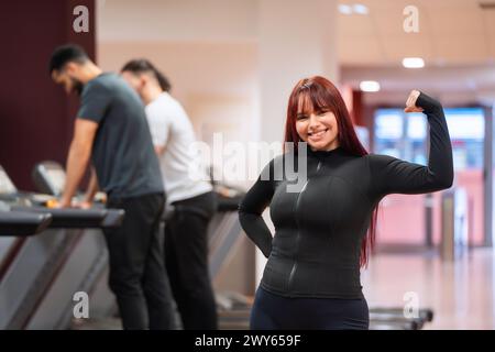 Lächelnde Frau in schwarzer Sportswear, die ihren Bizeps, Laufbänder und Menschen im Hintergrund bewegt. Stockfoto