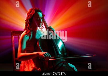 Porträt einer Cellistin, die klassische Melodien mit leuchtenden roten und violetten Bühnenlichtern hinter ihr auf der Bühne spielt. Stockfoto