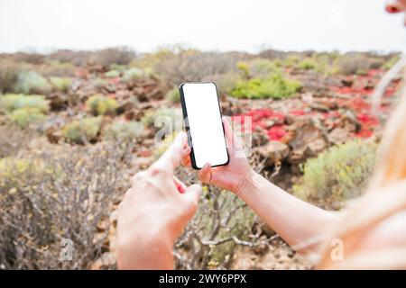 Eine Frau hält ein Handy in der Wüste. Sie zeigt auf den Bildschirm, macht vielleicht ein Foto oder sieht sich etwas am Telefon an. Stockfoto