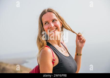 Eine Frau mit langen blonden Haaren lächelt und hält ihre Haare zurück. Sie trägt ein schwarzes Tanktop und eine goldene Uhr Stockfoto