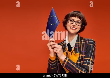 Glückliches junges College-Mädchen in Uniform und Brille, die EU-Flagge auf leuchtendem orangefarbenem Hintergrund hält Stockfoto