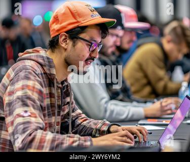 Ein Mann mit orangefarbenem Hut und Brille schreibt auf einem Laptop. Er ist von anderen Leuten umgeben, die auch Laptops benutzen. Die Szene scheint eine zu sein Stockfoto