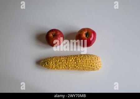 Gelb geschälte Maiskolben und zwei rote Äpfel bildeten Gesichtsemoji, zwei Augen und Mund auf weißem Tisch. Hausgemachtes echtes Bio-Gemüse nicht perfekt Stockfoto