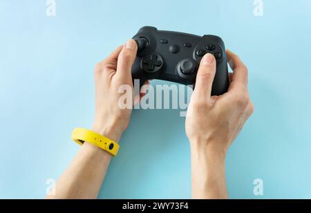 Zwei Hände greifen einen schwarzen Gamecontroller, der durch ein leuchtendes gelbes Armband auf hellblauem Hintergrund hervorgehoben wird. Zeit zum Spielen auf der Spielkonsole Stockfoto