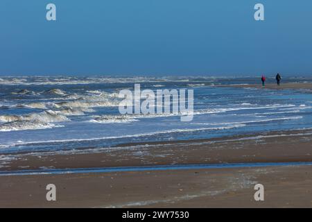 Egmond aan Zee, Niederlande - 17. März 2022: Wellen in der rauen Nordsee mit einigen Menschen, die in der Sonne am Ufer entlang laufen Stockfoto