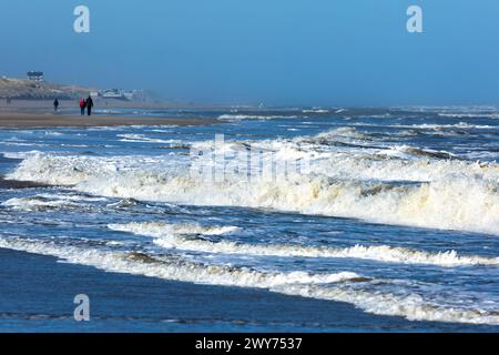 Egmond aan Zee, Niederlande - 17. März 2022: Wellen in der rauen Nordsee mit einigen Menschen, die in der Sonne am Ufer entlang laufen Stockfoto