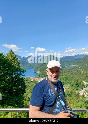 Reifer Mann mit Kamera über einer malerischen Klippe, lächelnd. Auf dem Berg bei Neuschwanstein, Bayern, Deutschland Stockfoto