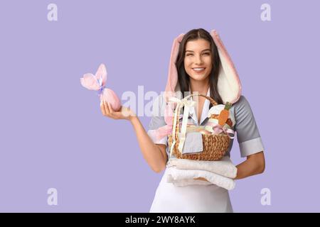 Glückliche Zimmermädchen in Hasenohren mit Osterkorb, Schokoladenei und Handtüchern auf lila Hintergrund Stockfoto