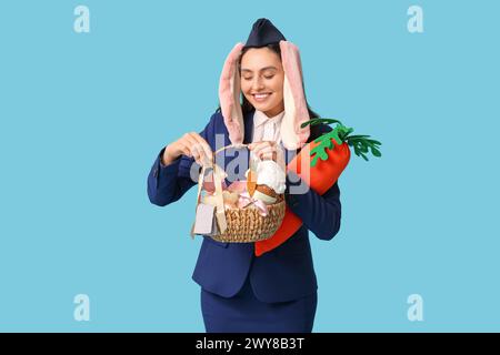 Glückliche Stewardess in Hasenohren mit Osterkorb und SpielzeugKarotte auf blauem Hintergrund Stockfoto