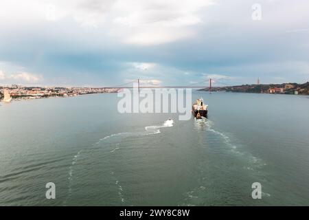 Luftdrohnenaufnahme eines Frachtschiffes, das im Fluss Tejo mit Ponte 25 de Abril im Hintergrund fährt. Der legendäre Ponte 25 de Abril überspannt den Hintergrund vor dem Hintergrund eines bewölkten Himmels. Stockfoto