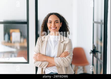 Attraktive, selbstbewusste, erfolgreiche hispanische oder brasilianische junge Frau mit lockigen Haaren, steht in einem modernen Bürobereich mit überkreuzten Armen, blickt und lächelt in die Kamera Stockfoto