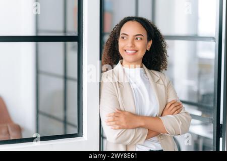 Foto einer selbstbewussten, wunderschönen, erfolgreichen hispanischen oder brasilianischen jungen Frau mit lockigem Haar, steht in einem modernen Büro mit überkreuzten Armen, blickt weg, träumt, lächelt freundlich Stockfoto