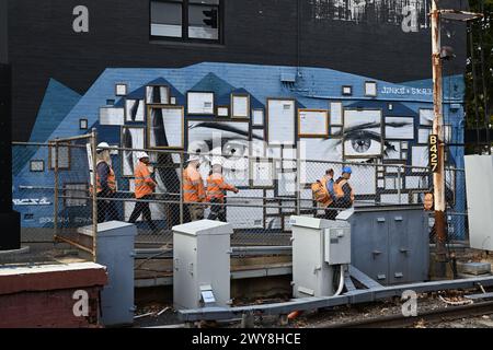 Eine Gruppe von Bahnarbeitern, die orangefarbene Warnwesten und Schutzhelme tragen, geht an einem großen Wandgemälde vorbei, wenn sie den Bahnhof Middle Brighton verlassen Stockfoto