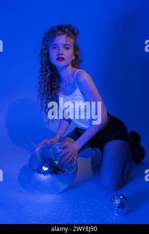 Eine junge Frau mit Disco-Ball posiert im Musikstil, während sie im Nachtclub feiert. Sinnlichkeit Frau kniend in blauen neonfarbenen Lichtern. Wunderschöne Frau Stockfoto
