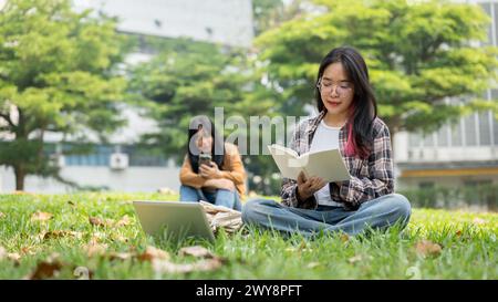 Eine fokussierte asiatische Schülerin liest ein Buch, während sie auf dem Gras sitzt, während eine andere Schülerin im Hintergrund ein Smartphone benutzt und dabei Outdo genießt Stockfoto