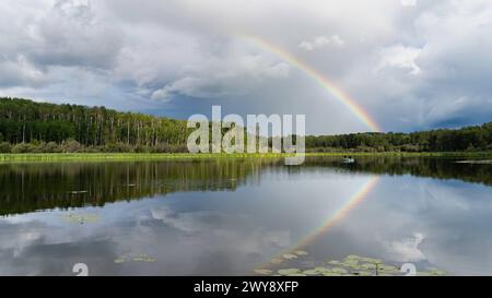 Ein Regenbogen spiegelt sich auf einem glasartigen See mit einem wolkengefüllten Himmel. Ein kleines Fischerboot schwimmt in der Ferne. Stockfoto