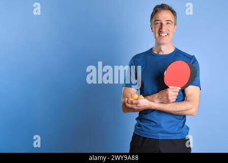 Lächelnder Mann in Sportbekleidung, der Tischtennispaddles und Bälle in den Händen auf blauem isoliertem Hintergrund zeigt. Stockfoto