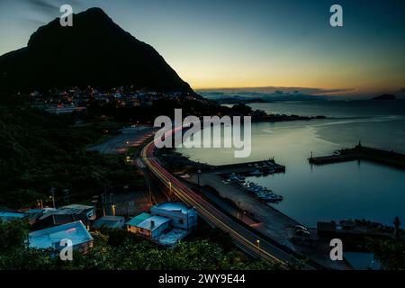 Leichte Autospuren entlang eines C-förmigen Abschnitts der Küstenstraße im Norden Taiwans begleiten die Landschaft mit Sonnenuntergang und Dorflichtern. Stockfoto