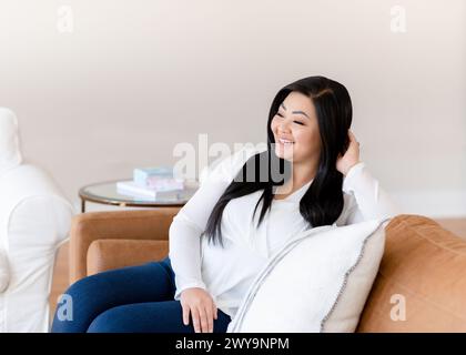 Asiatische Frau, die auf brauner Ledercouch sitzt und in der Ferne wegschaut Stockfoto