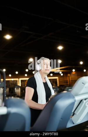 Fröhliche, hübsche ältere Frau in gemütlicher Kleidung, Training auf Laufband und Hören von Musik in Kopfhörern Stockfoto