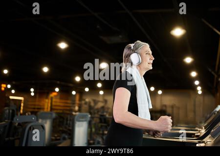 Positive, ziemlich reife Frau in gemütlicher Kleidung, Training auf Laufband und Musikhören in Kopfhörern Stockfoto