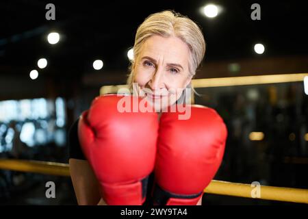 Ansprechende, fröhliche, reife Sportlerin mit Boxhandschuhen, die in die Kamera lächelt, während sie im Fitnessstudio klingelt Stockfoto