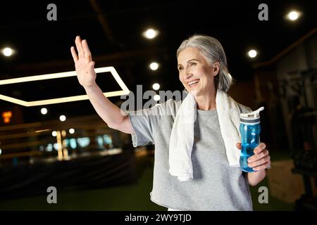 Ziemlich fröhlich reife Sportlerin mit Handtuch auf den Schultern und Wasserflasche in den Händen, die aktiv winkt Stockfoto