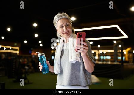 Fröhliche ältere grauhaarige Sportlerin in gemütlicher Kleidung, die mit einer Wasserflasche in den Händen auf das Telefon blickt Stockfoto