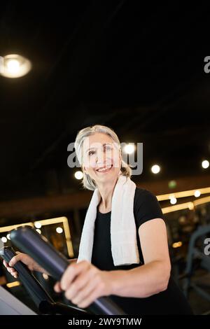 Ziemlich reife, fröhliche Frau mit grauem Haar und Handtuch auf den Schultern, die auf Crosstrainer trainiert Stockfoto