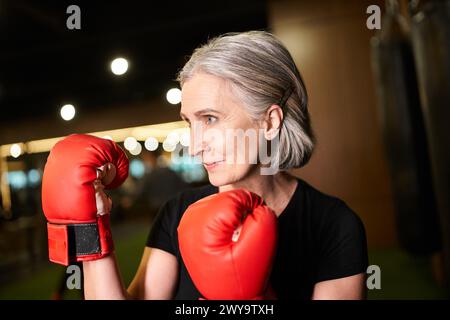 Sportlich gut aussehende Seniorin mit grauem Haar posiert mit Boxhandschuhen während des Trainings im Fitnessstudio Stockfoto
