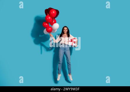 Großes Foto der guten Laune Frau gekleidetes Seidenhemd, das rote Blasen Geschenkbox in den Händen hält, isoliert auf blauem Farbhintergrund springt Stockfoto