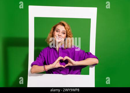 Foto-Porträt der schönen Dame halten sofortigen Bilderrahmen zeigen Herz Geste gekleidet stilvolle violette Kleidung isoliert auf grünem Farbhintergrund Stockfoto