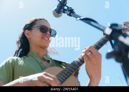 Junger lateinamerikanischer Mann mit langen Haaren und Sonnenbrille und grünem Hemd, Gitarrist Musiker mit guter Einstellung, der Rockmusik mit seiner akustischen Gestalt macht Stockfoto