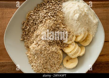 Nahaufnahme der Zubereitung eines tiefen Tellers mit Müsli-Zutaten, beginnend mit Haferflocken, Sonnenblumenkernen, Bananenscheiben, Proteinpulver. tischstatus Stockfoto