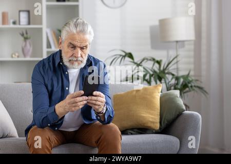 Ein reifer Mann mit grauen Haaren, der sich intensiv auf sein Smartphone in einem modernen Wohnzimmer konzentriert und ein Konzept der Technologienutzung bei älteren Menschen vermittelt. Stockfoto