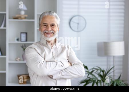 Porträt eines fröhlichen älteren Mannes, der zu Hause mit gekreuzten Armen steht und Selbstvertrauen und Positivität in einer komfortablen, stilvollen Umgebung ausstrahlt. Stockfoto