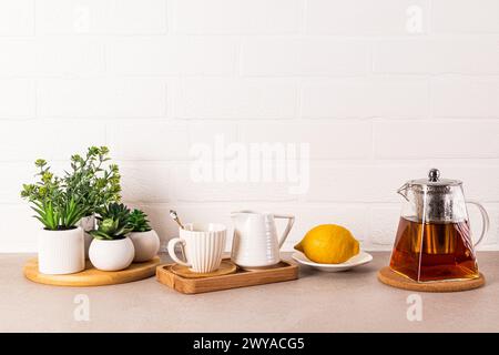 Schöner Küchenhintergrund mit Glas-Teekanne mit duftendem Tee, Tasse und Zitrone auf Tablett. Vorderansicht. Weiße Ziegelwand. Ein Kopierbereich Stockfoto
