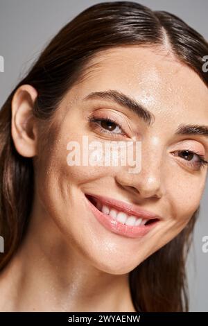 Eine junge kaukasische Frau mit brünetten Haaren und sauberer Haut, die hell lächelt, mit Wassertropfen, die ihr Gesicht bedecken. Stockfoto