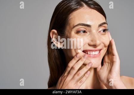 Eine junge kaukasische Frau mit brünetten Haaren lächelt warm und berührt sanft ihr Gesicht mit den Händen im Studio. Stockfoto