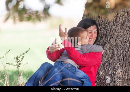 Berührender und liebevoller Moment, in dem der kleine Junge seine Mutter umarmt, während sie einen entspannenden Moment im Schatten einer Eiche genießen Stockfoto