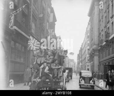 VE Day Scenes - Eine Straße in Richmond, Surrey. In der Mitte des Bildes ist ein Pferdewagen mit VE Day Revellers im Stehen und Sitzen zu sehen. Ein Mann sitzt auf der Rückseite des Wagens und hängt seine Beine über die Kante. Eine Frau sitzt auf den Schultern des Mannes und spricht mit einer anderen Frau, die im Wagen steht. Ein Mann, der nahe am Rand des Wagens stand, schwenkt mit einer kleinen Gewerkschaftsfahne. Rechts im Bild sehen die Personen, die auf dem Bürgersteig laufen, die Insassen des Wagens. Von einem Baum nach oben bis hinter die Kamera. Das Bild zeigt eine dichte Menschenmenge, die draußen versammelt ist Stockfoto