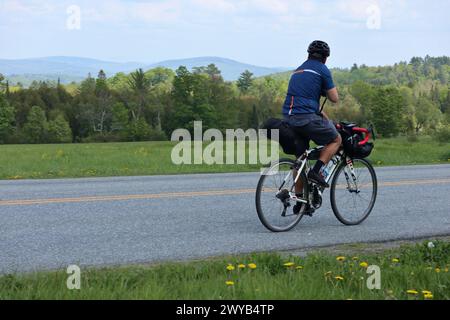 Mann, der auf einer Landstraße mit Bergen im Hintergrund fährt (kein Gesicht, wegschaut, Schotterrad mit Fahrradtaschen) Reisen, Touren, Stockfoto