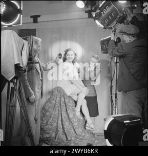 GEBURT Eines STARS: ALLTAG FÜR SCHAUSPIELERIN MURIEL PAVLOW, ENGLAND, 1945 – Schauspielerin Muriel Pavlow lächelt während eines Fotoshootings, wahrscheinlich in den Welwyn Garden Studios, Hertfordshire. Der Fotograf George Daley (links) bestimmt mit einem Lichtmesser die richtige Belichtung, während die Friseurin Elsie Bass Muriels Haar den letzten Schliff verleiht. Auf der rechten Seite ist ein Techniker zu sehen, der die Beleuchtung einstellt. , Stockfoto