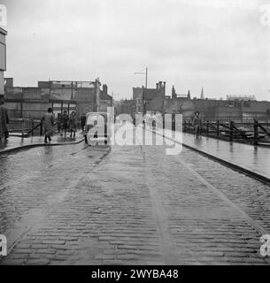 COVENTRY, FRÜHJAHR 1944: ALLTAGSLEBEN IN COVENTRY, WARWICKSHIRE, ENGLAND, Großbritannien, 1944 – westwärts entlang der Smithford Street in Coventry. Fußgänger sind auf beiden Straßenseiten zu sehen. Der regengetränkte Bürgersteig ist mit Holzgeländern gesäumt, die entlang leerer Räume verlaufen, in denen einst Geschäfte und Häuser standen. In der Ferne kann man den quadratischen Turm der St. John's (Bablake) Kirche sehen. , Stockfoto