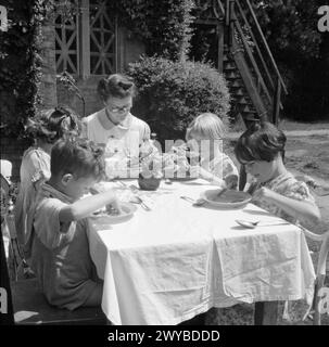 KINDERGARTEN: LEBEN IM ALTEN HERRENHAUS, WENDOVER, BUCKINGHAMSHIRE, ENGLAND, 1944 – Eine Krankenschwester setzt sich zusammen, um mit einigen der älteren Kinder an einem Tisch zu essen, der im sonnigen Garten des Kindergartens im alten Herrenhaus in Wendover aufgestellt ist. Der Originaltitel besagt, dass die Kindermahlzeiten, wann immer möglich, im Freien serviert werden. Eine kleine Vase mit Blumen ziert den Tisch. Im Hintergrund ist nur das Kreuzband an den Fenstern der Schule zu sehen, um Explosionsschäden zu vermeiden. , Stockfoto