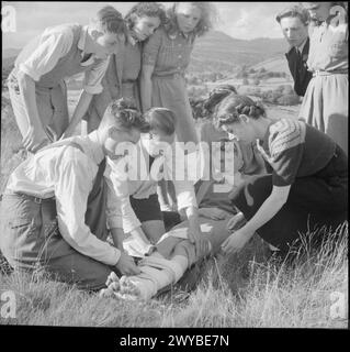 KOOPERATIVE JUGENDSOMMERSCHULEN: BILDUNG UND UNTERHALTUNG, CRAIG SCHOOL, WINDERMERE, ENGLAND, VEREINIGTES KÖNIGREICH, 1943: Eine Gruppe junger Menschen, die die Co-operative Union Summer School an der Craig School am Lake Windermere besuchen, üben erste Hilfe bei einem Spaziergang auf den Fjells. Ein Mädchen sitzt auf dem Gras, während zwei Jungen ihre Beine zusammenbinden, eines wird als Schiene für das andere, gebrochenes Bein benutzt. Sie wird auch von zwei Mädchen besucht, von denen eines ihren Rücken unterstützt, während der Rest der Gruppe zusieht. Die erste-Hilfe-Kurse an der Summer School in Windermere wurden von Herrn F Kay von der St. John's Ambulance Brigade geleitet. , Stockfoto