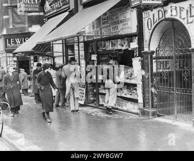 LONDON IM FRÜHJAHR 1941: ALLTAGSLEBEN IN LONDON, ENGLAND – Männer und Frauen schauen sich die Schlagzeilen an, wenn sie an einem Zeitungskiosk in der Londoner Fleet Street vorbeikommen. Die Taverne Old Bell ist ebenfalls rechts auf dem Foto zu sehen. , Stockfoto