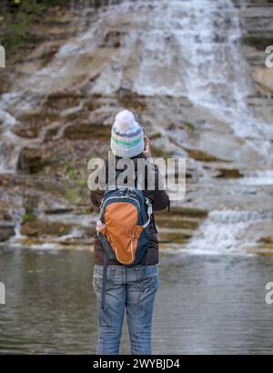 Frau, die ein Foto von einem Wasserfall im Herbst mit wechselnden Blättern auf ihrem Handy macht (Wanderer mit Rucksack und Jacke, Hut macht Handyfoto o Stockfoto