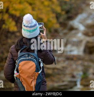 Frau, die ein Foto von einem Wasserfall im Herbst mit wechselnden Blättern auf ihrem Handy macht (Wanderer mit Rucksack und Jacke, Hut macht Handyfoto o Stockfoto