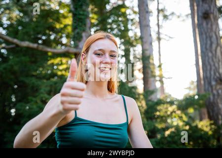 Eine Frau lächelt und gibt die Daumen hoch. Sie trägt ein grünes Tanktop und steht im Wald Stockfoto